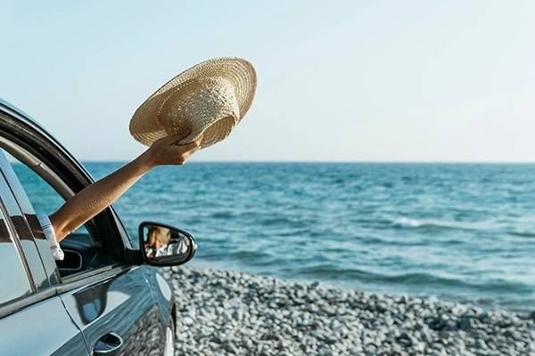 Mulher com o braço para fora do carro segurando um chapéu de praia