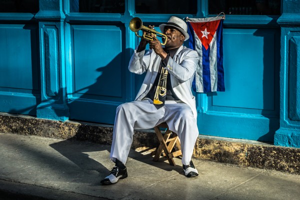 Hombre cubano sentado en la acera tocando la trompeta con una bandera de Cuba de fondo en La Habana.