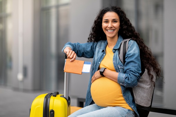 Mujer embarazada feliz posando para la foto con pasaporte, boletos de avión y maleta de viaje en la mano.