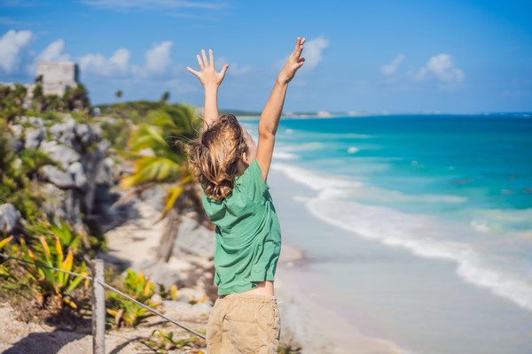 Niño alzando los brazos en el borde del acantilado de la ciudad amurallada de Tulum, Riviera Maya, México, con el mar Caribe de fondo.