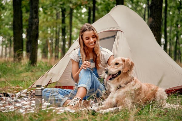 Mujer afuera de una tienda de campaña en un bosque o camping con un perro.