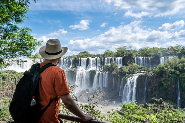Hombre con mochila y sombrero admirando las Cataratas del Iguazú desde una pasarela en un día soleado, Parque Nacional Iguazú, Argentina.