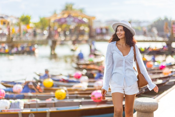 Mujer caucásica vestida de blanco caminando en un muelle junto al río en H?i An, Vietnam, mientras observa los botes con una sonrisa.
