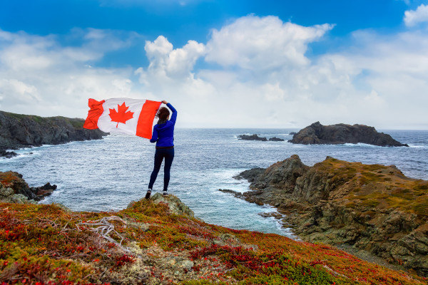 Mujer de espaldas sosteniendo bandera de Canadá al viento sobre costa rocosa en el océano Atlántico en Twillingate, Terranova.