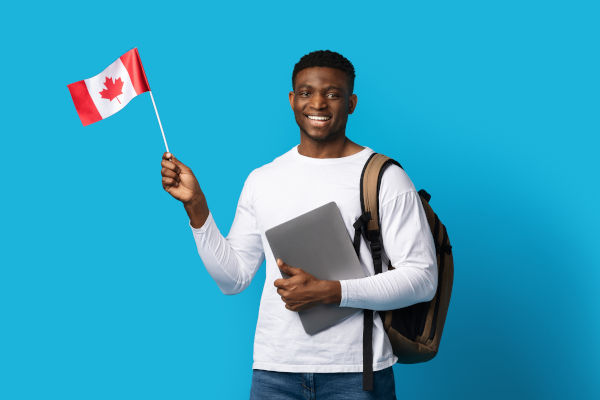 Joven sonriente con mochila de viaje sosteniendo una bandera canadiense en una mano y una laptop en la otra.