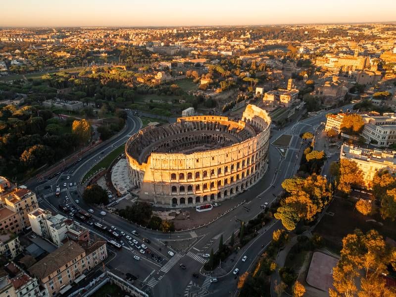 Vista panorâmica do Coliseu em Roma com turistas caminhando pela praça em um dia ensolarado na Itália.