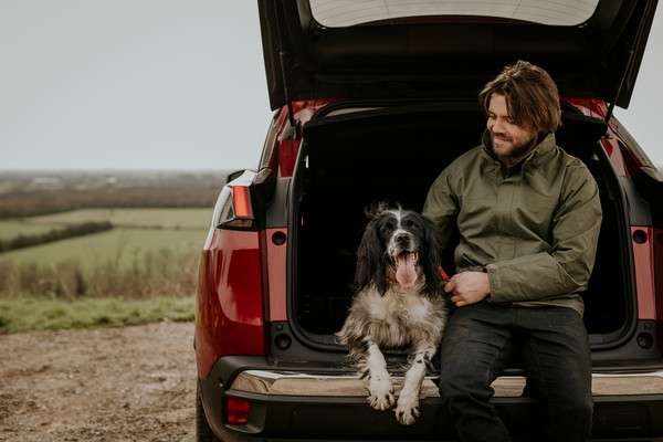 Hombre sentado en el maletero del coche con su perro con un paisaje rural de fondo.