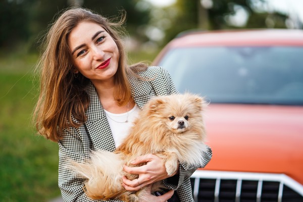 Mujer parada en frente de su auto, sonriendo ligeramente a la cámara con un perro pomeranio en brazos.