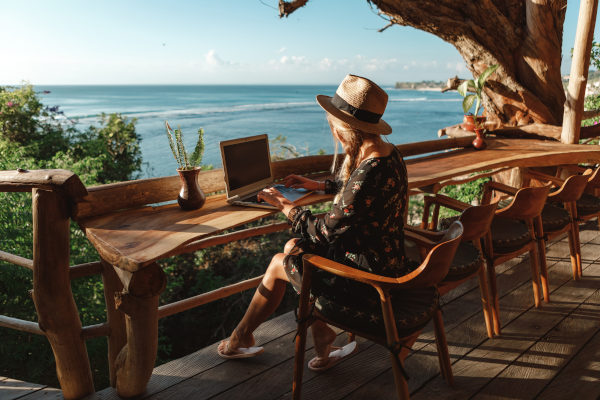 Mujer usando un ordenador portátil en una terraza de una cafetería al aire libre con vistas al mar.