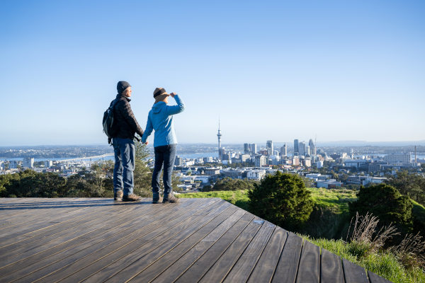 Pareja de pie en el malecón de la cumbre del Monte Edén, con el Sky Tower y el Puente del Puerto de Auckland en la distancia, Nueva Zelanda.