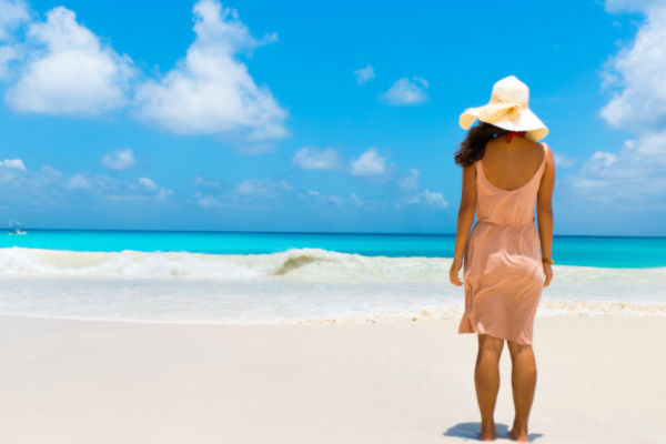 Mujer joven parada frente al mar de Aruba, con un sombrero y vestido rosa.