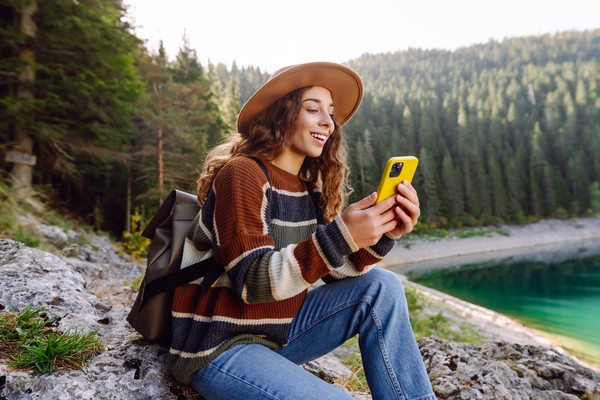 Joven viajera con mochila y ropa de abrigo sentada en una roca junto a un hermoso lago, sonriendo y usando su teléfono móvil.