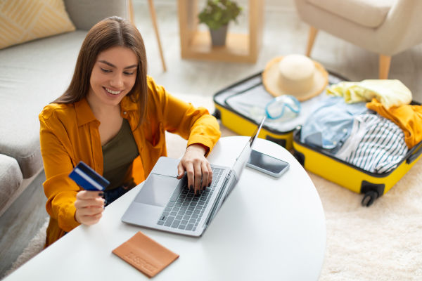 Mujer sonriente sosteniendo una tarjeta mientras usa una notebook, con una valija de viaje de fondo.