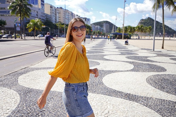 Mujer turista sonriente con gafas de sol caminando por la calzada de Copacabana en un día soleado en Río de Janeiro, Brasil, con calles, palmeras y arena de fondo.