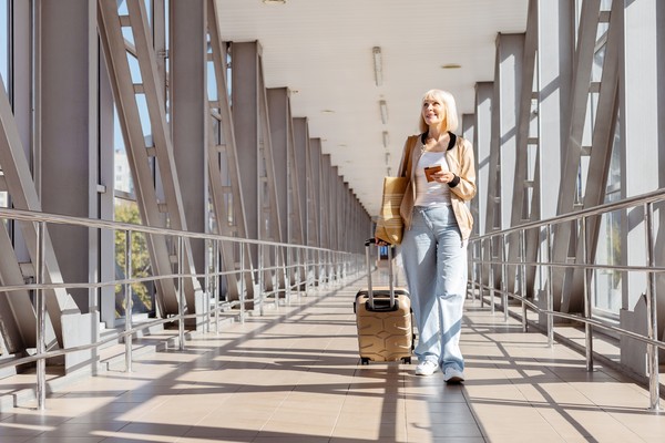Mujer adulta sonriente y tranquila con pasaporte en mano caminando con valija por un aeropuerto, lista para embarcar.