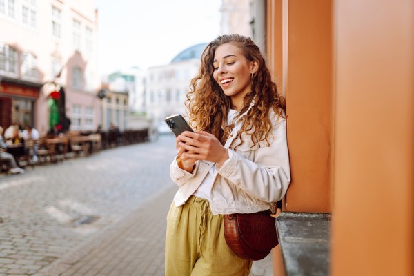 Mujer feliz usando el teléfono contra una pared en una calle empedrada.