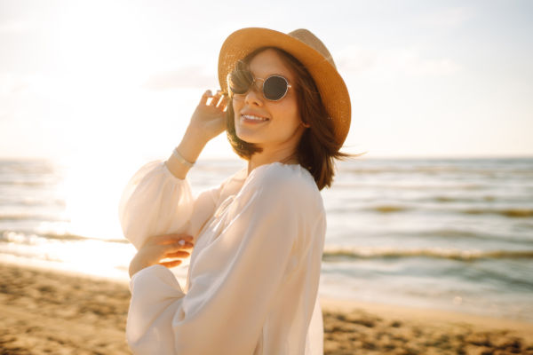 Mujer sonriente con sombrero y gafas de sol disfrutando del atardecer en una playa de Camboriú, Brasil.