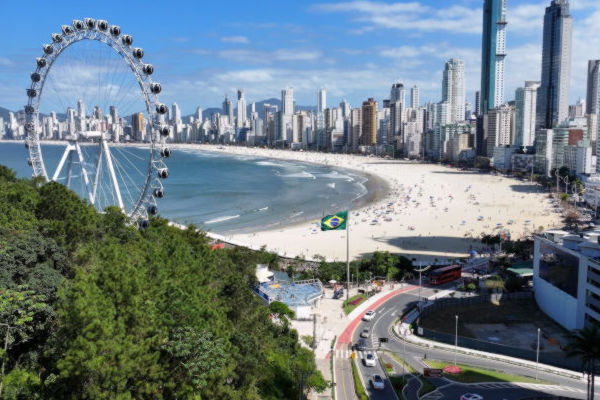 Vista panorámica de la playa central de Balneário Camboriú con la noria gigante, edificios altos y bandera de Brasil en primer plano.