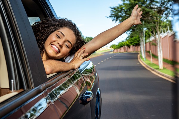 Mujer joven feliz saludando por la ventanilla de un auto en movimiento por la carretera.