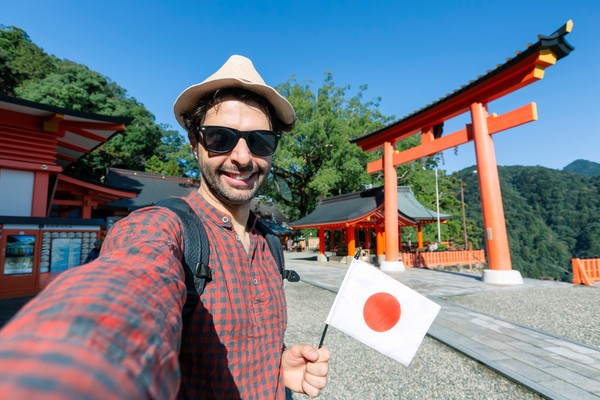 Joven turista masculino con una banderita de Japón tomándose una selfie junto a un tori en un día soleado en Japón.