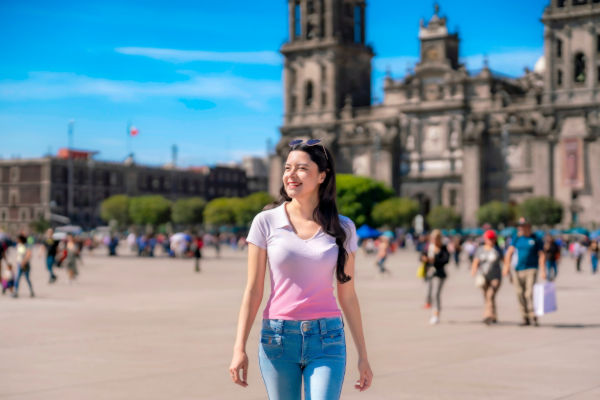 Mujer sonriente caminando por el Zócalo de Ciudad de México, con la catedral de fondo y turistas alrededor.