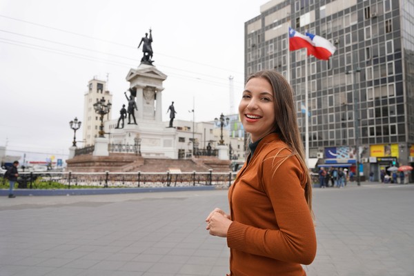Mujer turista sonriente en la Playa Sotomayor de Valparaíso con monumento y bandera de Chile de fondo en un día nublado.