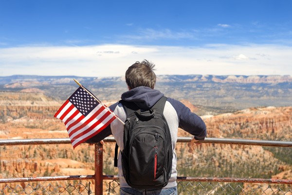 Joven de espaldas contemplando un paisaje árido con una mochila de la cual asoma una pequeña bandera de Estados Unidos.