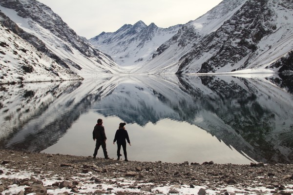 Dos turistas de pie en la orilla de la Laguna Inca en Valparaíso, Chile, admirando las montañas nevadas de fondo en un día nublado
