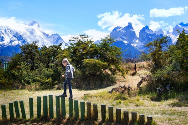 Niño jugando sobre una valla de troncos con un paisaje de montaña de fondo en el Parque Nacional Torres del Paine, Patagonia chilena.