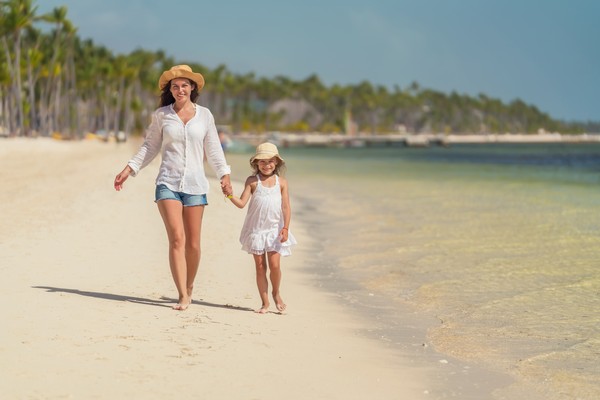 Madre e hija con sombreros caminando de la mano descalzas por una playa en República Dominicana con vegetación de fondo