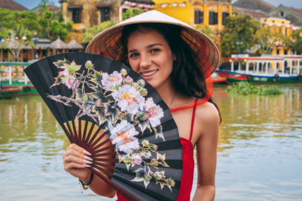 Mujer sonriente con vestido rojo y sombrero tradicional vietnamita, junto a un río en Hoi An.