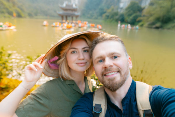 Pareja de turistas tomando una selfie frente a un lago en Vietnam, con sombrero tradicional y fondo de botes.