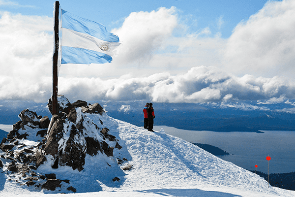 Imagem de duas pessoas com roupa de neve e a bandeira da Argentina hasteada em Bariloche.