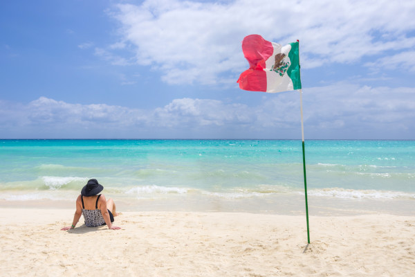 Mujer con sombrero sentada en una playa caribeña en un día soleado con una bandera de México clavada en la arena a poca distancia de ella.