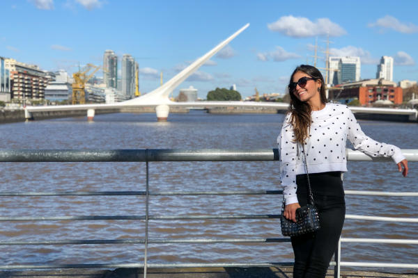 Mujer joven con camiseta a lunares y lentes de sol posando en una barandilla en Puerto Madero, con el Puente de la Mujer de fondo, sobre el Río de la Plata