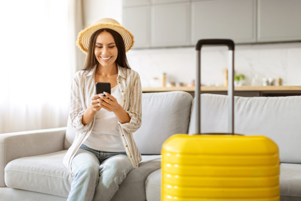 Mujer con sombrero de turista sentada en un sillón sonriendo con la vista en su smartphone y una maleta al lado