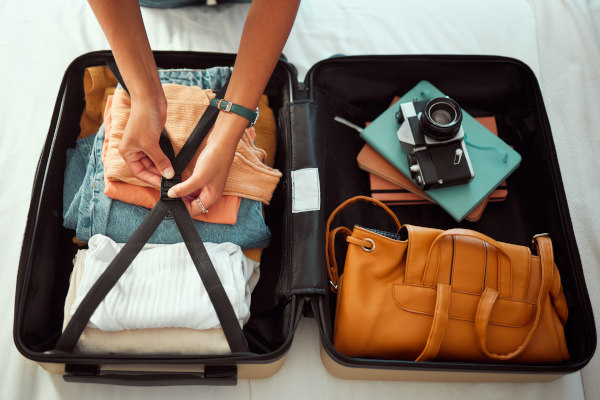 Manos ajustando ropa en una maleta abierta con prendas, cartera de mano, libretas y cámara fotográfica