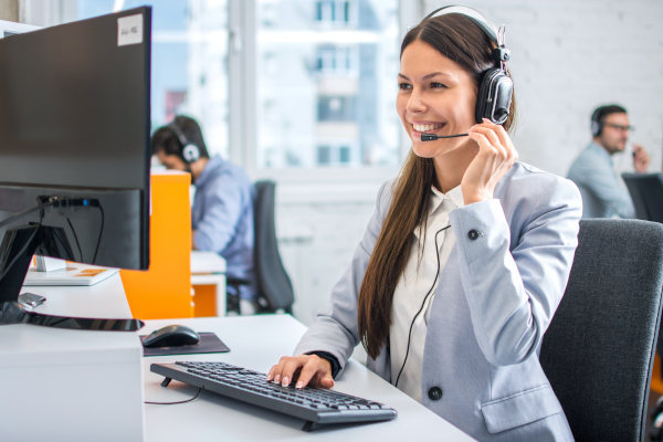 Personal femenino de central de atención al cliente sonriendo frente a una computadora mientras ajusta el micrófono de su headset.