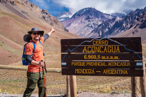 Hombre con equipo de trekking señalando hacia arriba a un lado del letrero del Cerro Aconcagua que indica la altura del mismo.