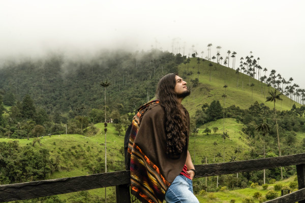 Joven latino de pelo largo con una ruana andina tejida apoyado en una valla y disfrutando del paisaje en el Eje Cafetero, Colombia.
