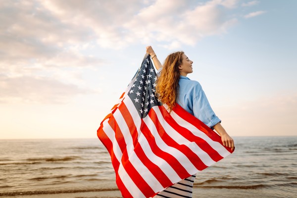 Mujer sonriente sosteniendo bandera de Estados Unidos en una playa desconocida al atardecer.