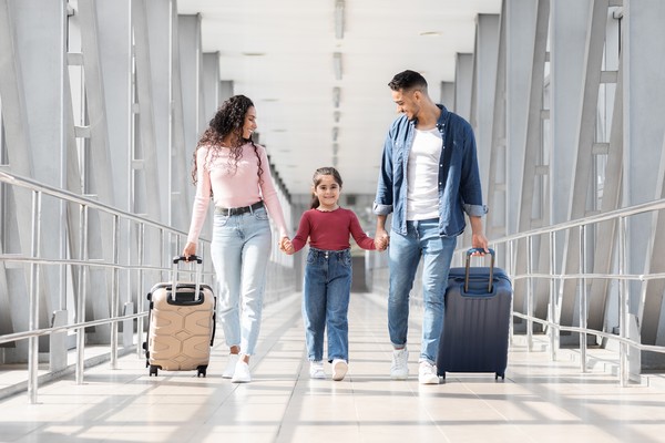 Pareja feliz con hija de la mano cargando maletas por la terminal de un aeropuerto.