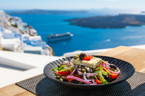 Plato con ensalada griega con el mar de fondo y construcciones tradicionales en la isla de Santorini, Grecia.