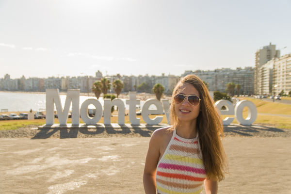 Mujer turista con cabello largo y lentes de sol posando con una sonrisa frente al Letrero de Montevideo, cerca de la Rambla, Uruguay