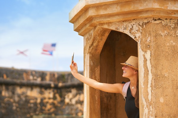 Mujer turista tomando fotografías con smartphone en una estructura antigua del Viejo San Juan, Puerto Rico, con bandera de Puerto Rico y Estados Unidos de fondo.