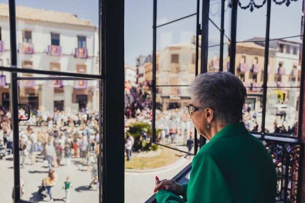 Mujer mayor mirando por la ventana una procesión de Semana Santa que ocurre en la calle.