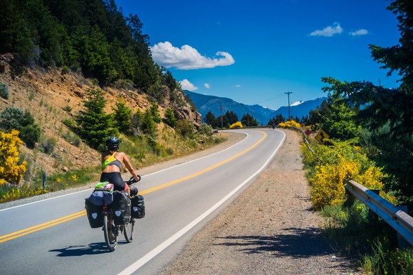 Mujer en bicicleta en una ruta patagónica con montañas a un lado y de fondo.