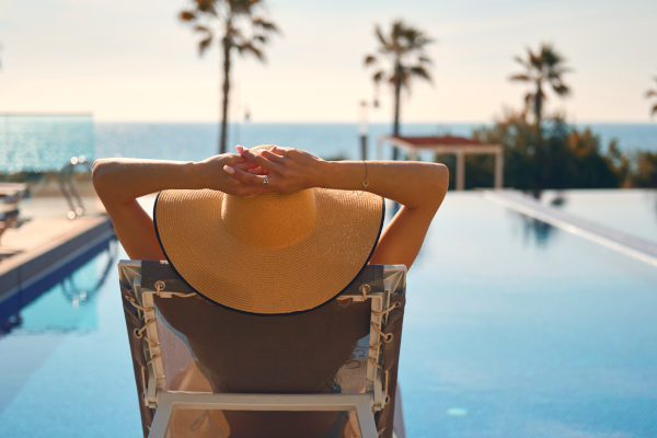 Turista con sombrero semiacostada en una reposera frente a la piscina de un resort en un destino tropical con palmeras de fondo.