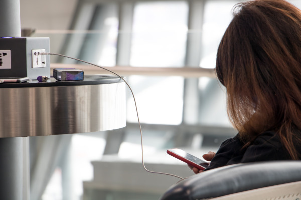 Mujer usando el celular mientras lo carga en un cargador público de celulares en un aeropuerto mediante cable USB.