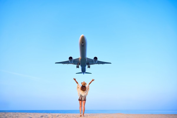 Mujer en una playa con los brazos levantados mientras un avión pasa por el cielo en ascenso por encima de ella.
